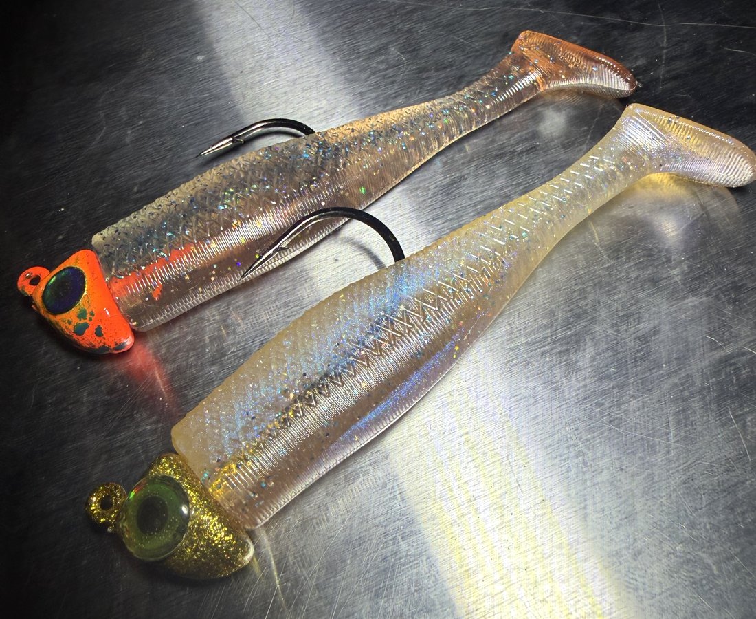Two rigged swimbaits on orange and gold jig heads under a shaft of light