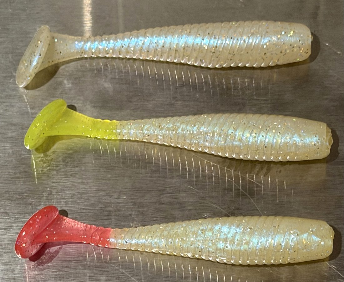 Three pearl swimbait bodies with clear, chartreuse and red tails on a steel bench